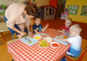 Chłopcy siedzą przy stoliku i odciskają swoje dłonie pomalowane w farbie na kartce. Pani Kinga stoi za jednym z chłopców i pomaga mu malować pędzelkiem umoczonym w farbie liść.